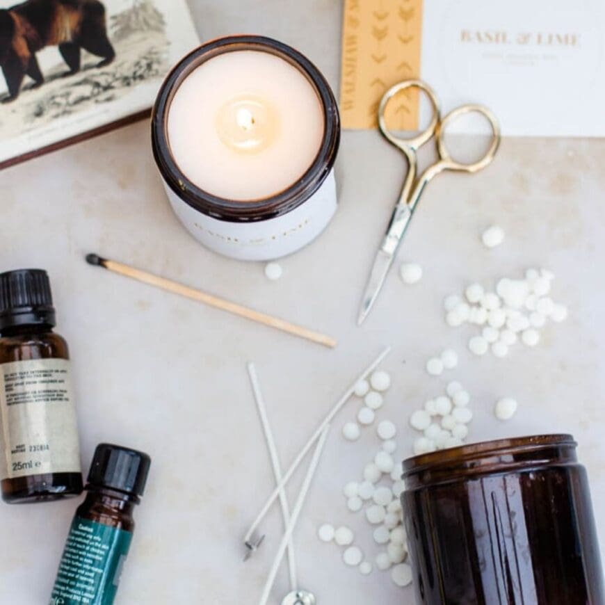 The ingredients for making candles. A lit candle in  the center with a small pair of scissors, a match, two bottles of essential oils, three wicks and a scattering of soy wax pellets