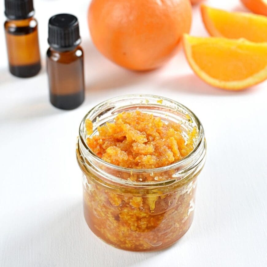 A jar of homemade salt scrub  with two bottles of essential oil in the background and orange segments