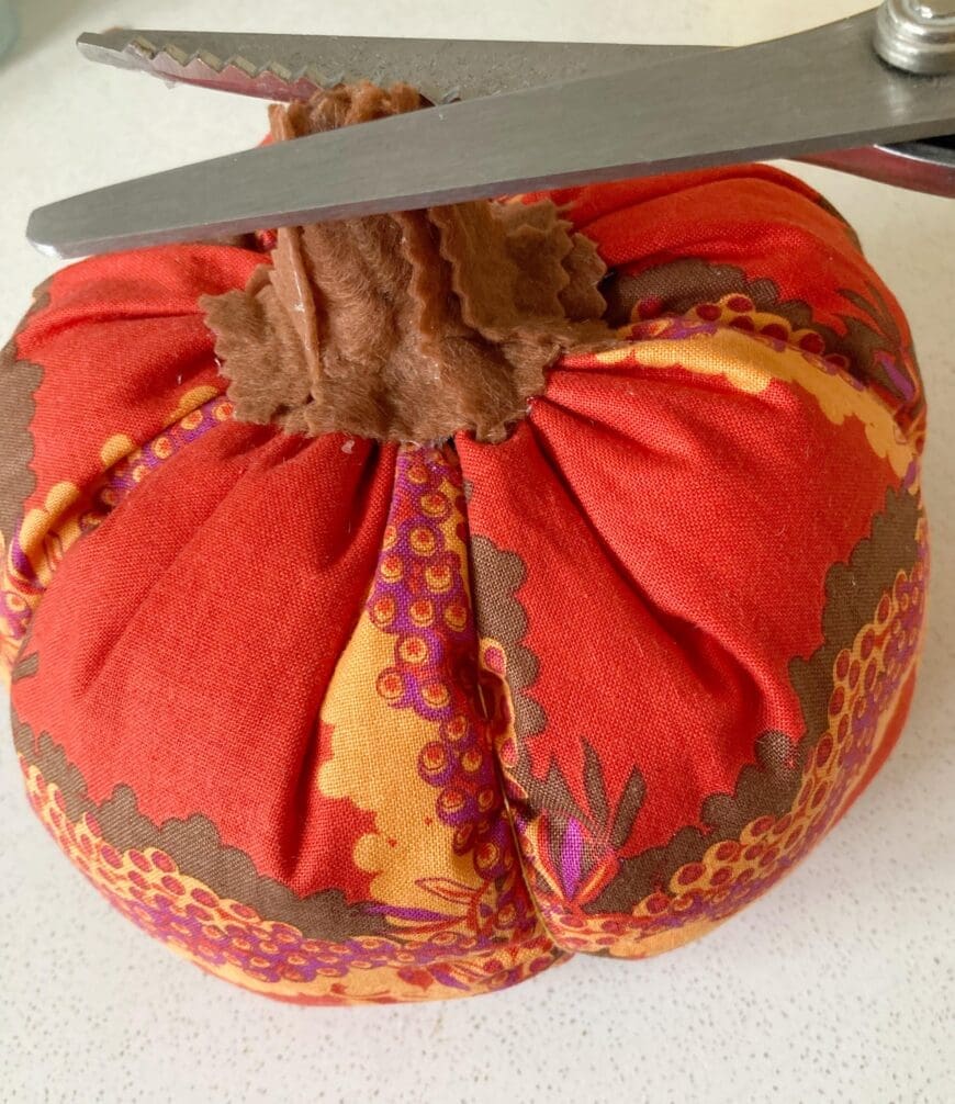 A Red fabric pumpkin with brown felt stalk being trimmed with pinking scissors 