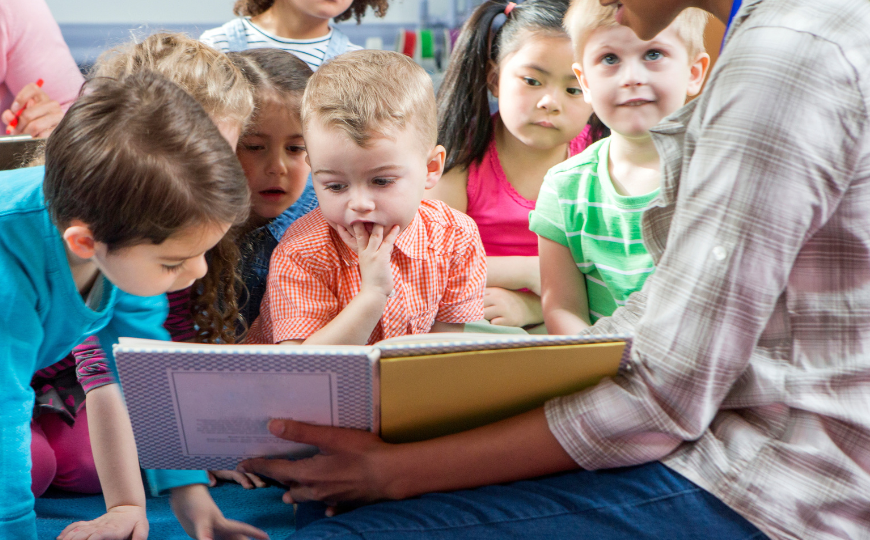 Children attentively listening to a teacher read all sat around her on the floor